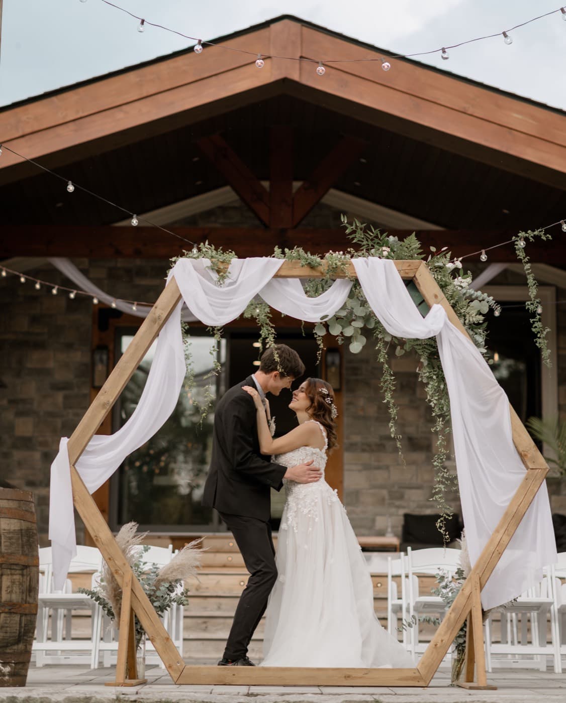 A couple stand forehead to forehead inside the draped hexagonal arbour, the stone pavilion and string lights behind them