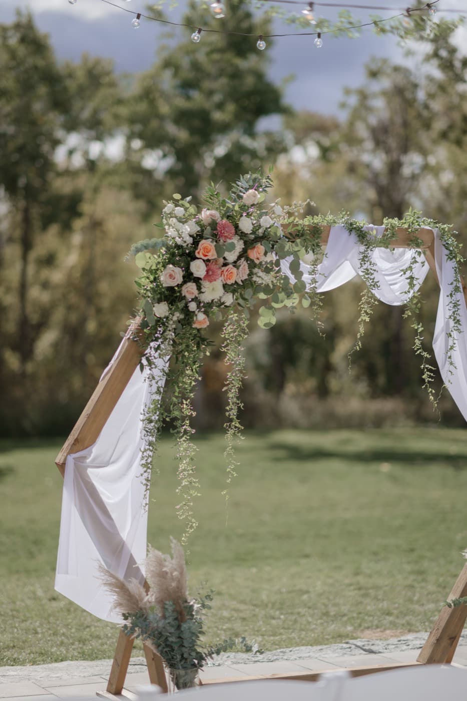 The corner of the ceremony arbour close up, banked with peach and cream roses, eucalyptus and trailing greenery over white drapery
