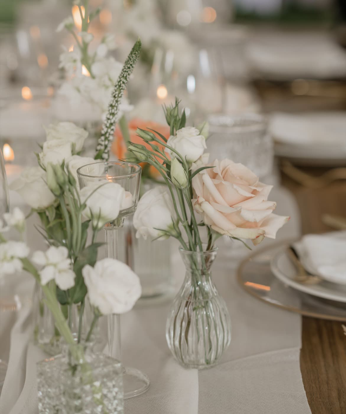 Small glass bud vases of white and blush roses running down a candlelit reception table