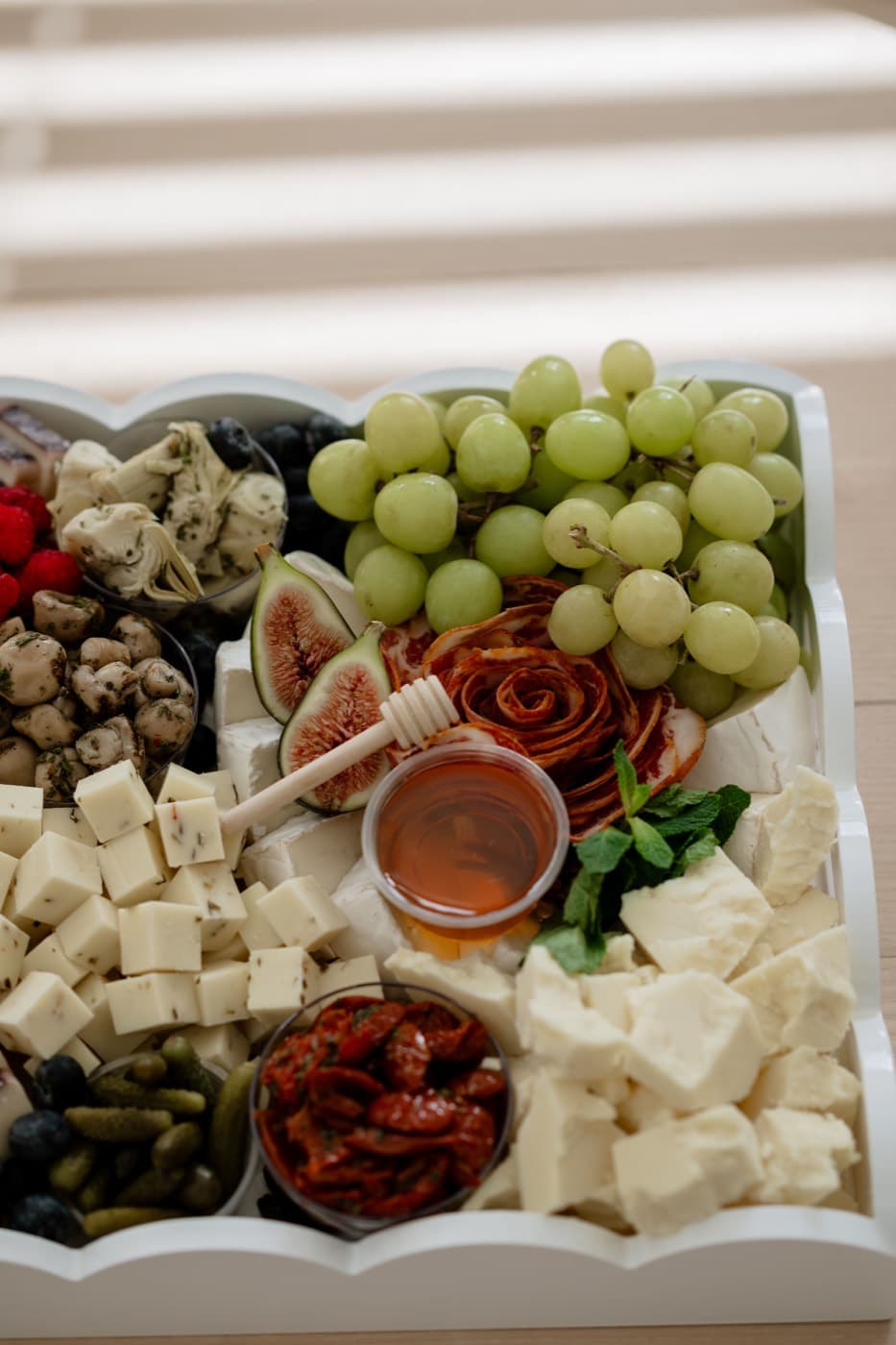 A white scalloped tray of cubed cheeses, green grapes, figs, marinated mushrooms, cornichons and a pot of honey