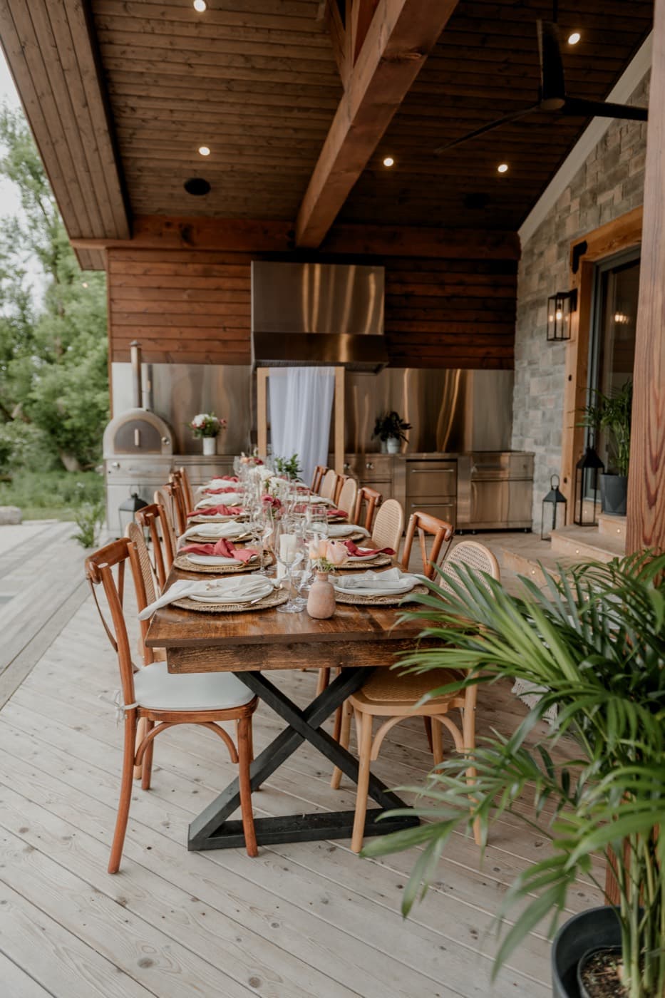 The covered pavilion with its timber ceiling and stone wall, a long harvest table set for dinner beside the outdoor kitchen