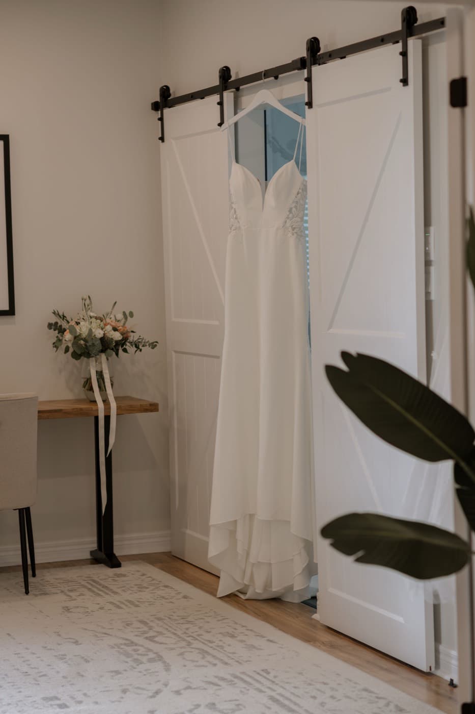 A wedding dress hanging against white barn doors, the bouquet resting on a wooden console table beside it