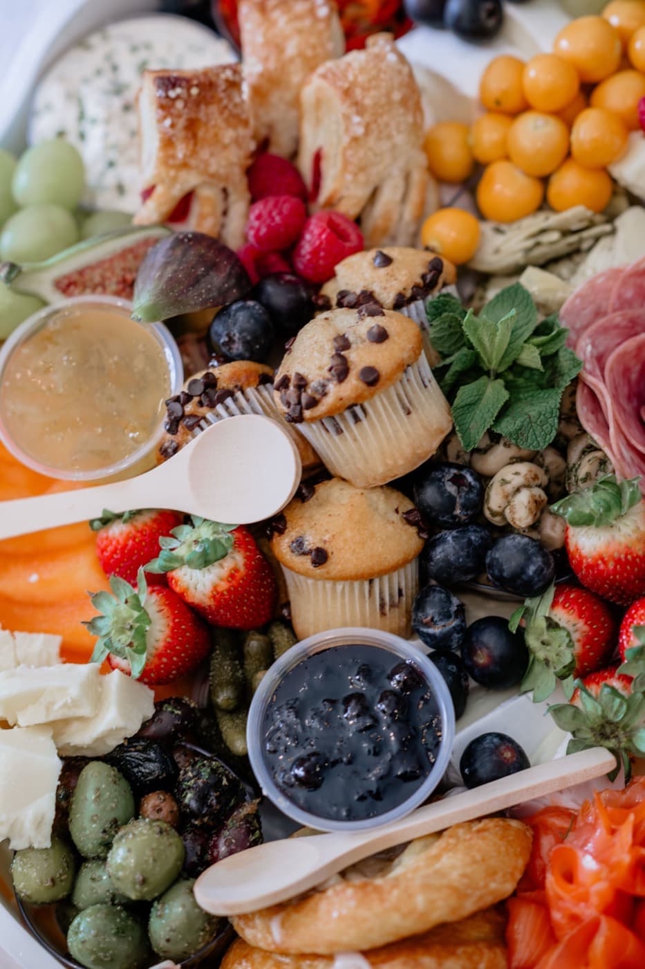 A grazing platter close up, filled with pastries, muffins, strawberries, blueberries, figs, olives, cured meats and small pots of preserves