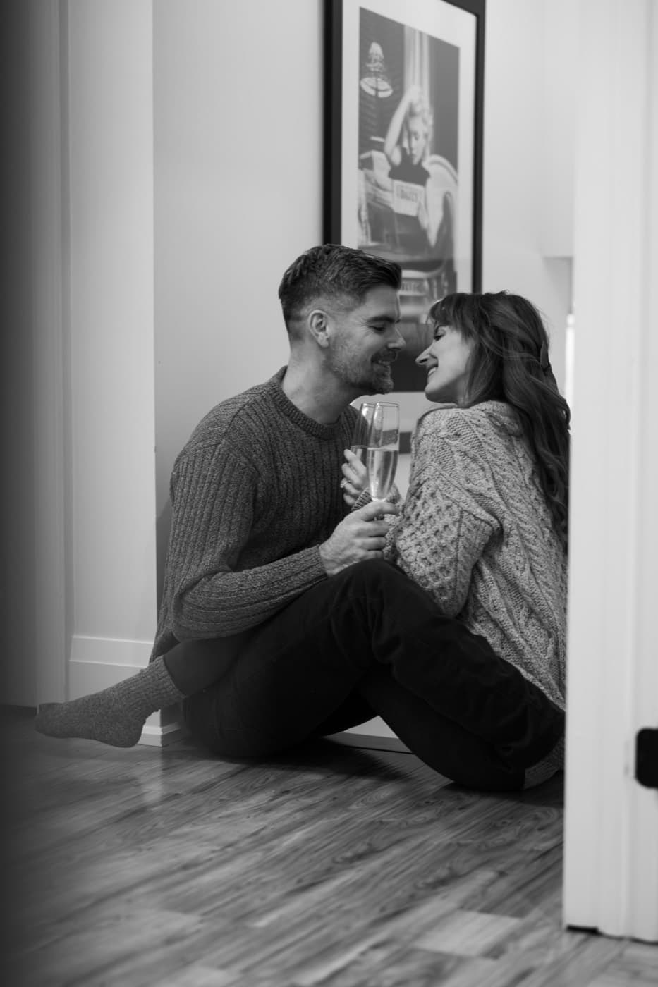 A couple sit together on the floor indoors, sharing a quiet toast