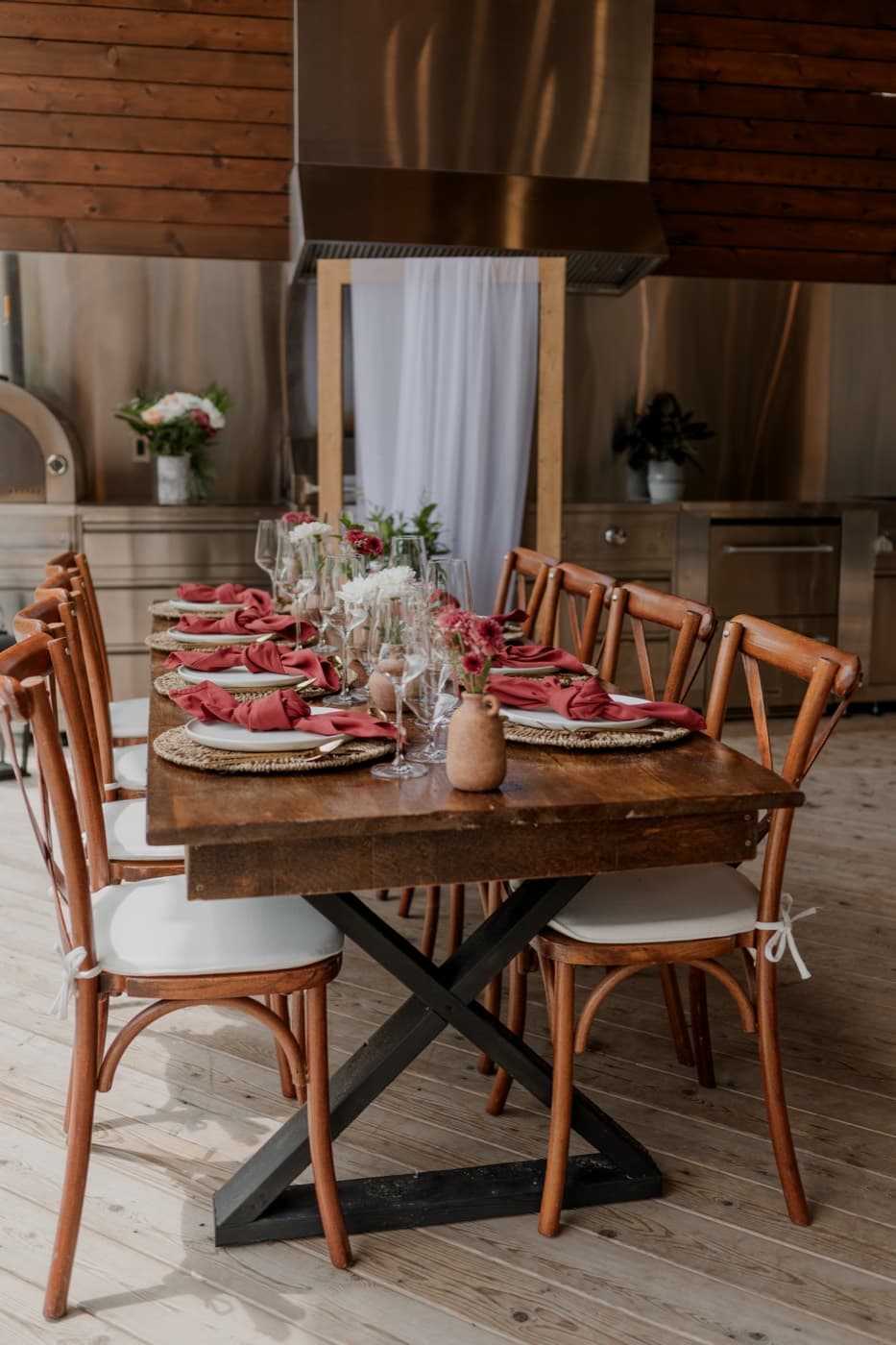 A long harvest table with cross-back chairs and rust napkins standing on the pale wood deck of the outdoor kitchen