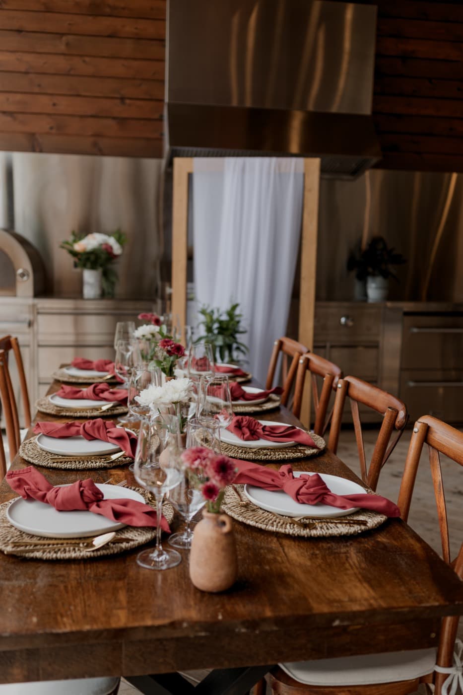 A long wooden table set with rust napkins and rattan chargers in the outdoor kitchen, the pizza oven behind it