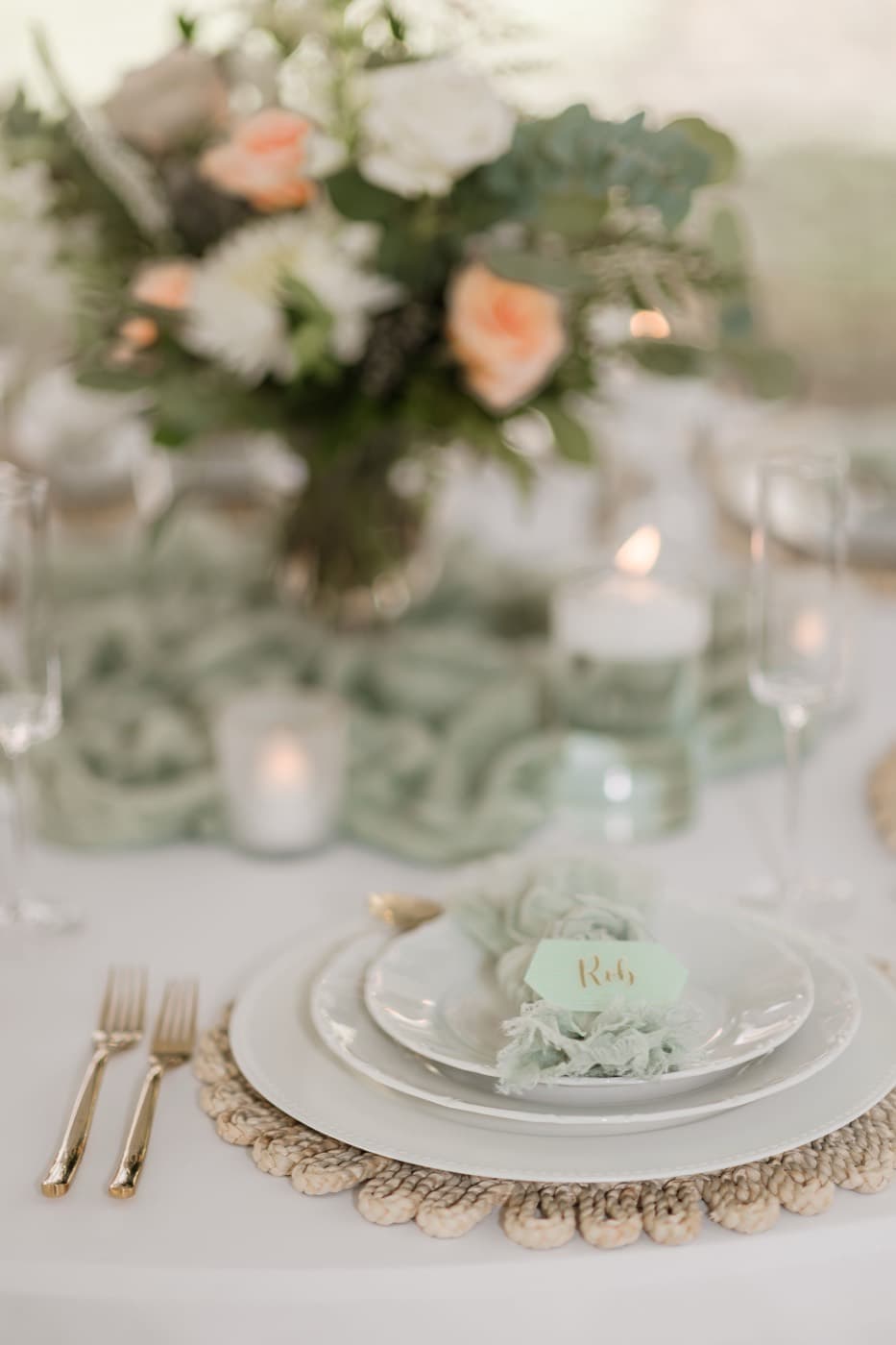 A place setting close up with a rattan charger, sage napkin and hand-lettered place card, the centrepiece soft behind it