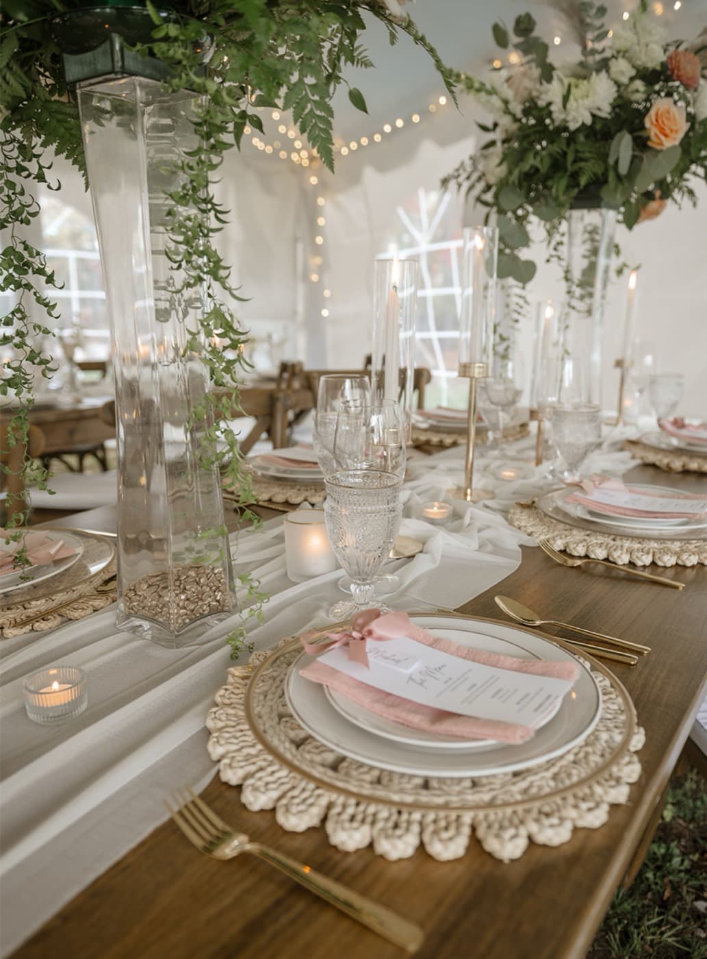 A long reception table set with gold cutlery, blush napkins, candles and trailing greenery under string lights