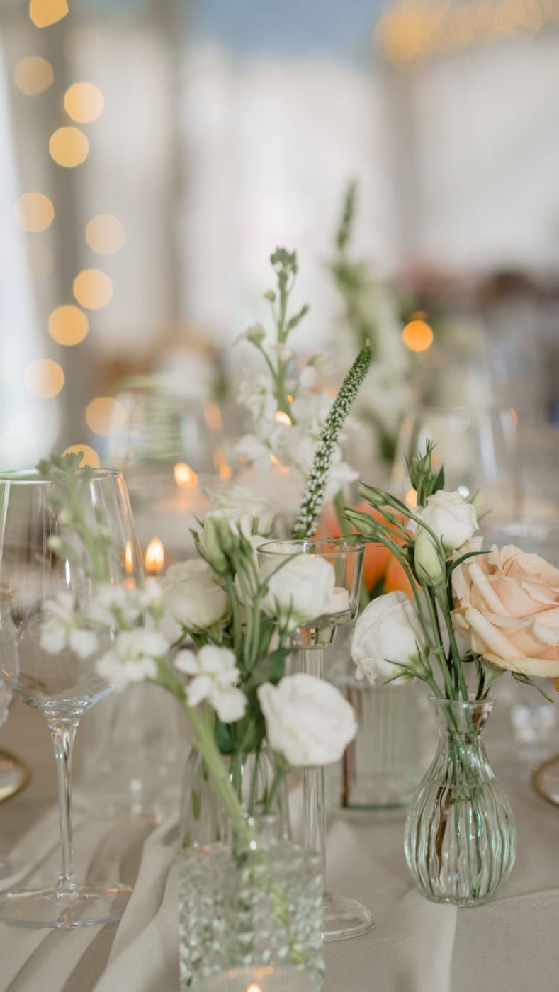 Bud vases of white and peach roses among lit candles, the string lights of the tent blurred behind them