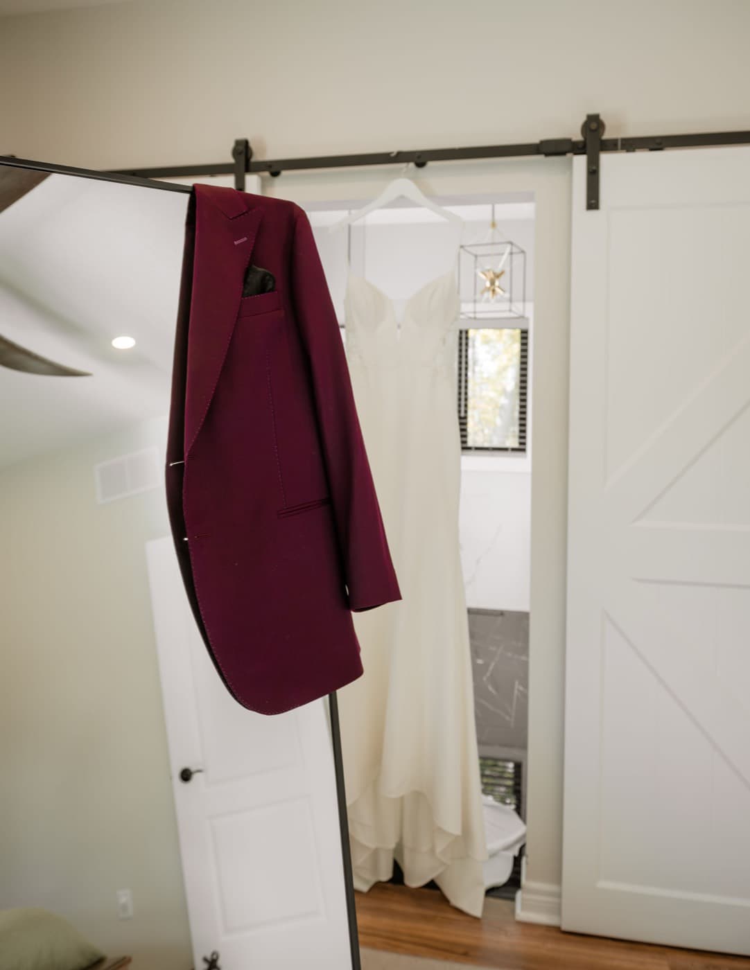 A burgundy suit jacket hanging from a barn-door track, the wedding dress hanging in the doorway behind it