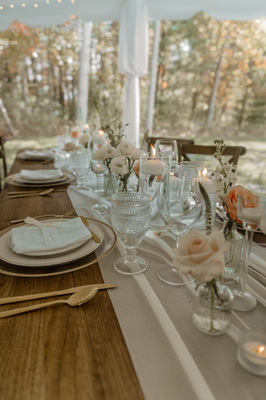 A reception table with floating candles and bud vases, autumn woods visible through the open side of the tent