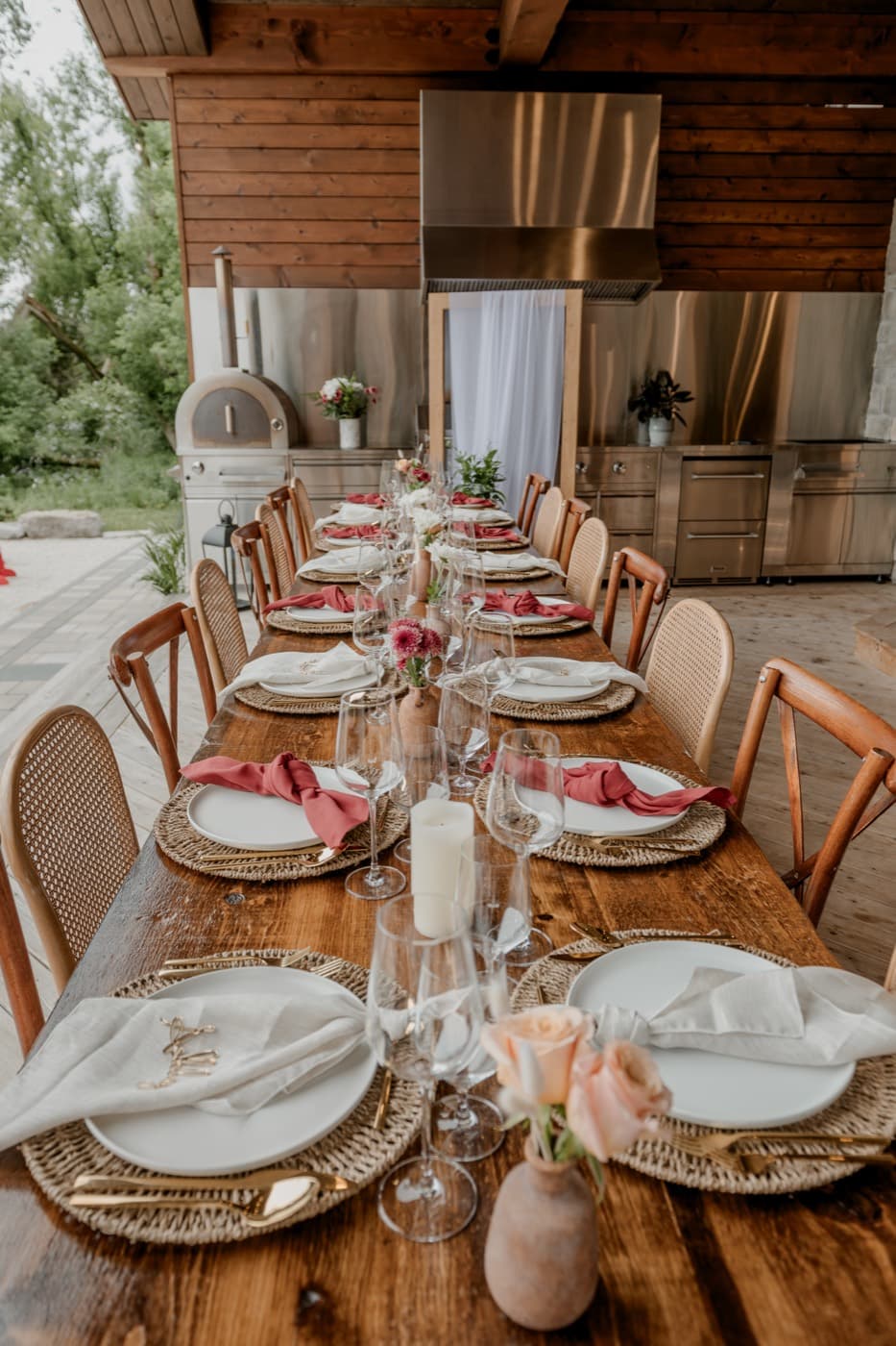 The set table seen down its full length, rust and cream napkins on rattan chargers and terracotta vases of roses, the pizza oven behind