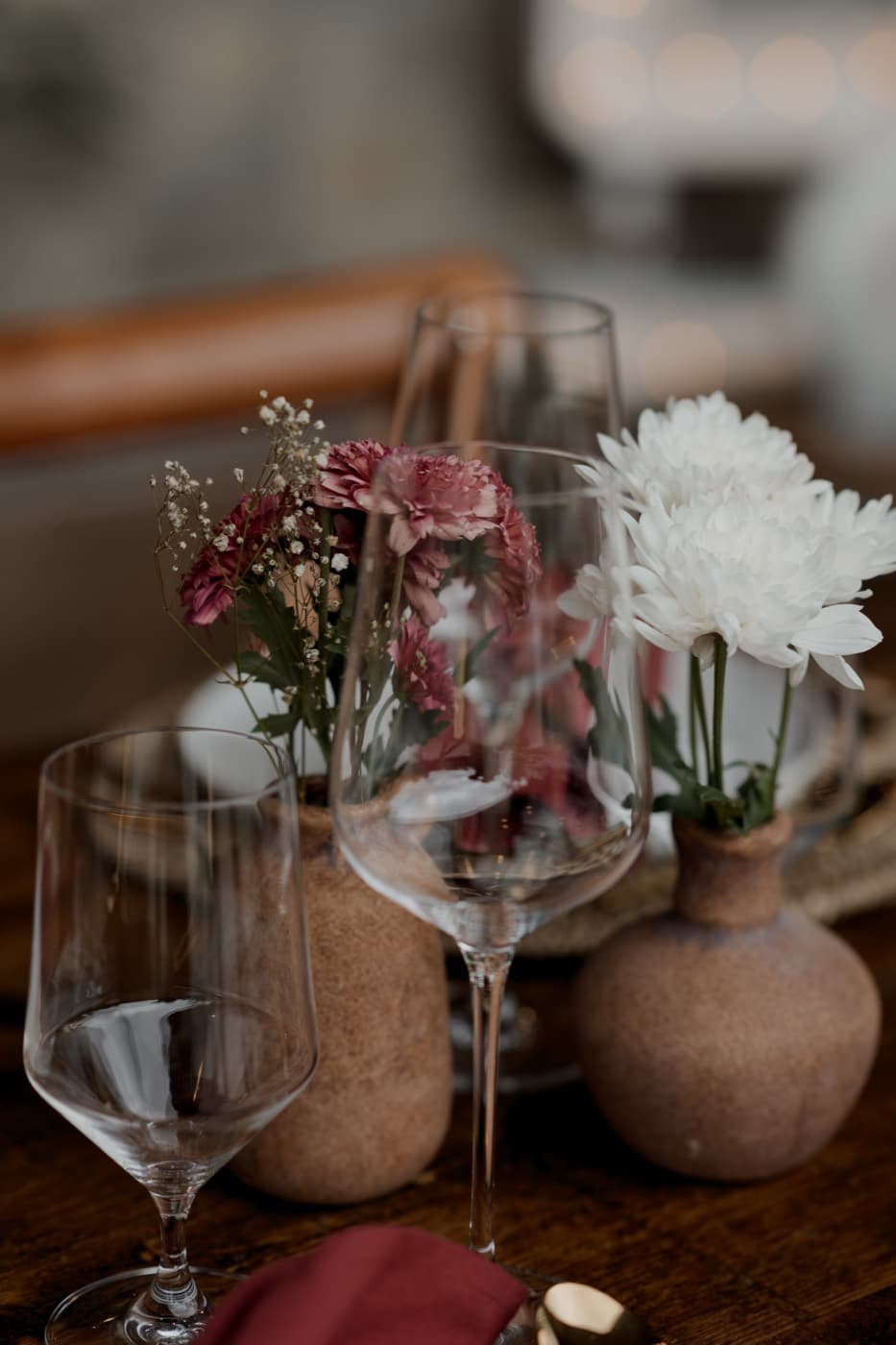 Terracotta bud vases holding a white chrysanthemum and pink carnations between wine glasses on a wooden table