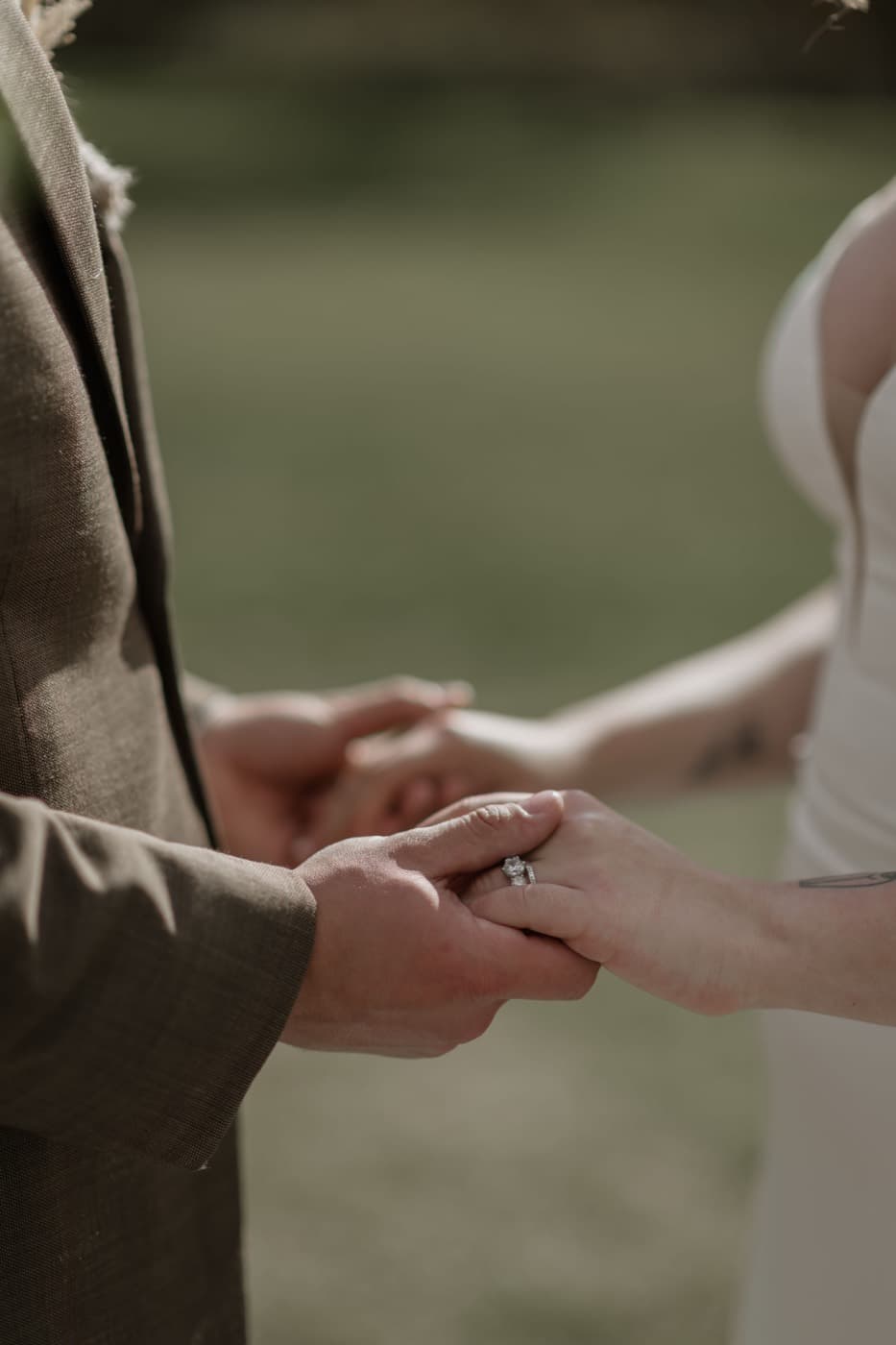 A couple hold hands during their vows, close enough to see the ring, the groom in a brown suit and the bride in white